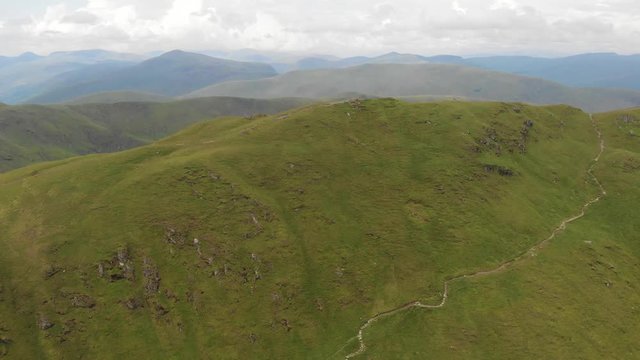 An aerial forward footage of a grassy mountain rocky summit with small pond and mountain range in the background under a cloudy sky
