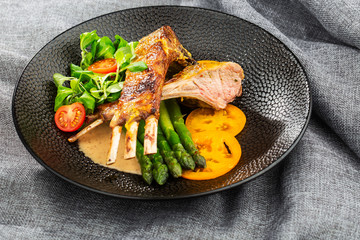 Roasted rack of lamb served with asparagus, yellow tomatoes and lamb sauce. Loin lamb Served in black stone plate with fork and knife. Gray background. Tuna salad. Top view from above horizontal.