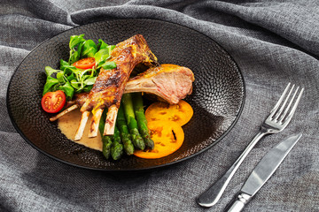 Roasted rack of lamb served with asparagus, yellow tomatoes and lamb sauce. Loin lamb Served in black stone plate with fork and knife. Gray background. Tuna salad. Top view from above horizontal.