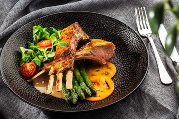Roasted rack of lamb served with asparagus, yellow tomatoes and lamb sauce. Loin lamb Served in black stone plate with fork and knife. Gray background. Tuna salad. Top view from above horizontal.