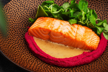 Fried fish salmon fillet with pink humus garnish. One piece of baked salmon grilled lime, tomatoes and salade on a brown plate . Gray background. Horizontal photo.