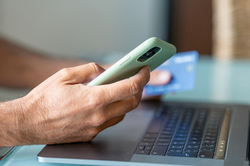 Man doing online shopping at home. Detail on hands holding smartphone and credit card with modern laptop keyboard on background