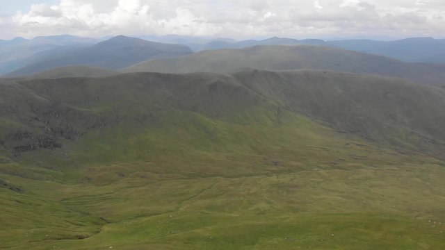 An aerial backward footage of a grassy mountain rocky summit with small pond and mountain range in the background under a cloudy sky