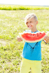 A blond boy holds a watermelon slice in his hands. The boy is eating a watermelon