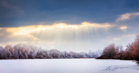 Sunbeams penetrate a dark cloud over a snow-covered field and trees. Winter landscape at sunset_