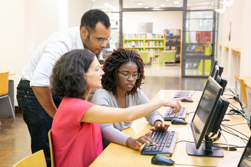 Instructor helping students in computer class. Man and women sitting and standing at desk, using desktop, pointing at monitor and talking. Training course concept