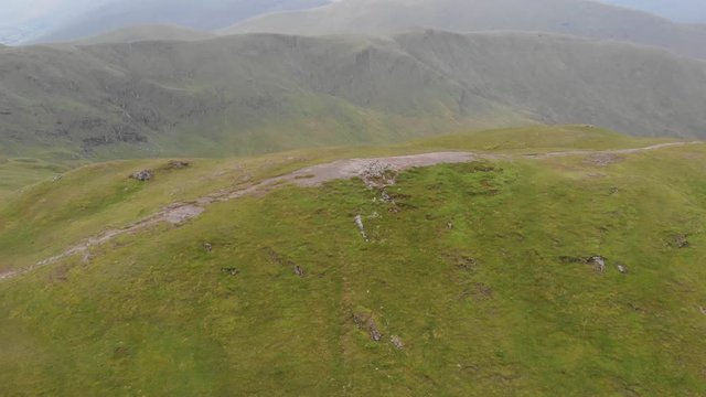 An aerial circular footage of a grassy mountain rocky summit with small ponds and mountain range in the background under a cloudy sky