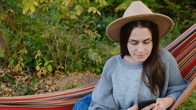 Young woman hanging on hammock distracted by social media or game applications, dressed grey sweater and hat, slow motion. Social media influencer people or content maker concept in relax at nature