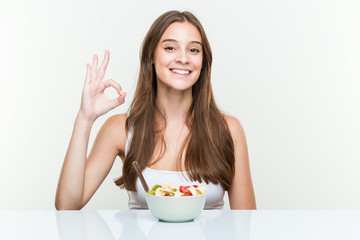 Young caucasian woman eating fruit bowl cheerful and confident showing ok gesture.