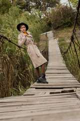 Fototapeta premium Beautiful girl on a suspension bridge. Woman in a black hat and a beige trench coat. Pretty girl on the suspension bridge. Fashionable autumn woman. Girl in black leather boots.