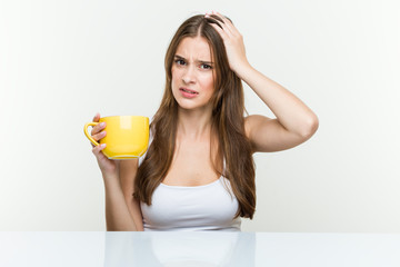 Young caucasian woman holding a cup being shocked, she has remembered important meeting.