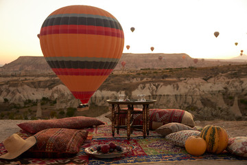Obraz premium big colourful balloon rises in the blue sky above a sandy hill on the ground there is a Turkish carpet with pillows, a plate of fruit and two pumpkins on it and a table in the middle of the carpet