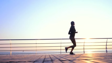 Athletic young girl jogging in the morning by sea wooden embankment. Silhouette of girl in sports costume. Beautiful sunlight. Healthy lifestyle concept
