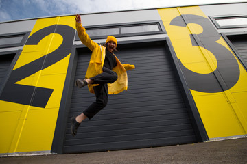 young caucasian woman wearing a yellow hat and a yellow raincoat jumping on a city street, positive...