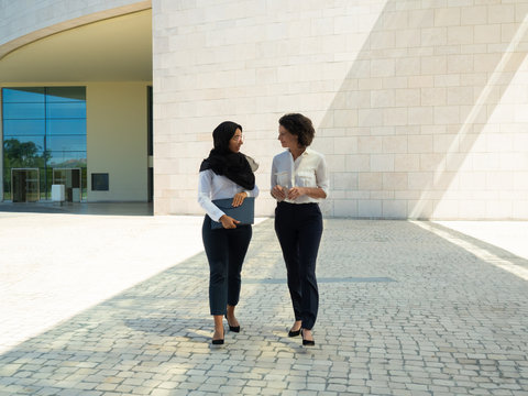 Diverse female business colleagues chatting, while going outside. Young Muslim businesswoman walking from office building, holding folder and talking to colleague. Communication concept