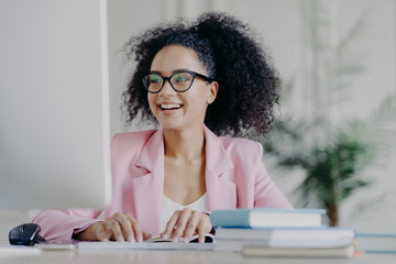 Photo of glad curly professional female manager focused into computer screen, smiles broadly, has...