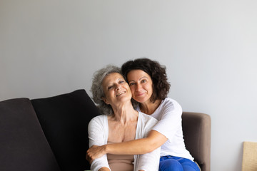 Happy cheerful middle aged woman hugging senior lady at home. Adult daughter sitting on couch and...