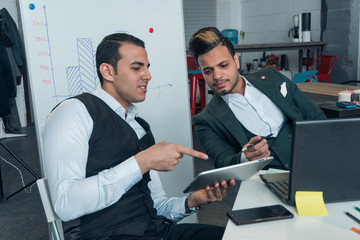 Two young men in business suits are negotiating and looking at business profit charts in a tablet