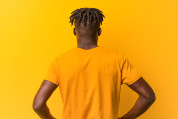 Young black man wearing rastas over yellow background from behind, looking back.