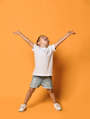 Cute kid boy in white t-shirt and blue shorts is posing with his hands up wide spread on yellow with copy space above