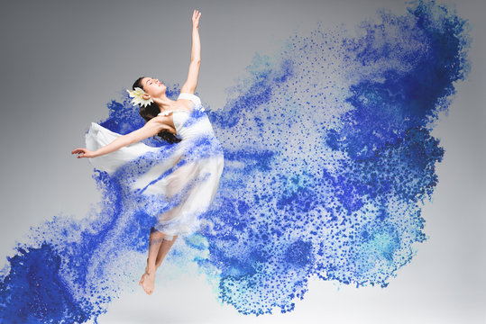 young ballerina in white dress dancing in blue paint splashes on grey background