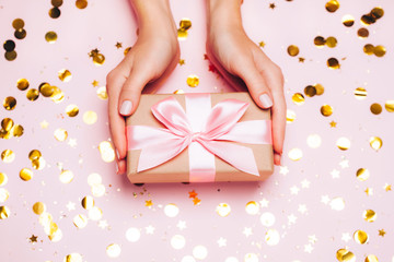 Female hands holding gift present box on festive confetti background. Top view, flat lay. Christmas greeting card.