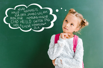 pensive kid standing with backpack near chalkboard with greetings lettering on green