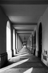 Monochrome. Arcades with shadows of columns on the pavement - covered walkway enclosed by a line of arches outside the building on a sunny day. 