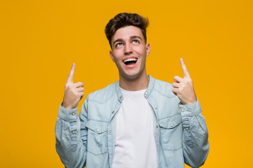Young handsome student wearing a denim shirt indicates with both fore fingers up showing a blank...