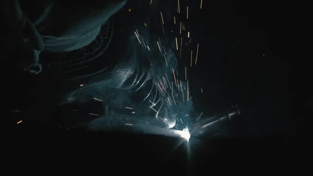 Forge workshop. Smithy. Worker in a welding hood helmet welds a part by electric welding. Sparks are reflected in the protective screen.