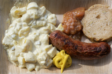 roast sausage with potato salad, mustard and bread n a bamboo plate, fast food to go on a street festival, top view from above