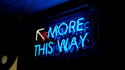 A bright blue fluorescent 'More This Way' neon sign with a yellow arrow pointing upwards. The neon sign illuminates its surroundings making this a key feature to the growing establishment