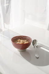 Morning oatmeal in red bowl and modern spoon on the white table with shadows from transparent curtain