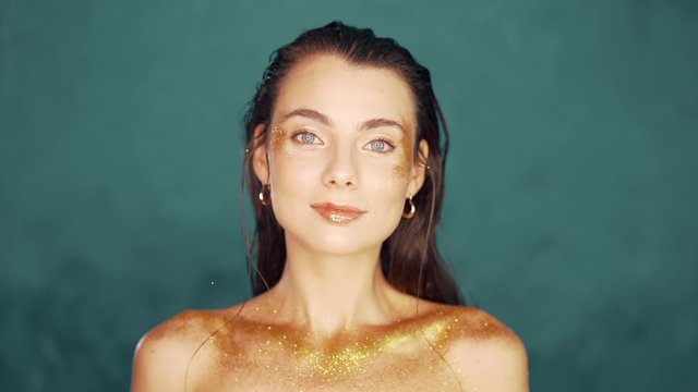 Perfect naked girl covered with gold paint and shimmering sparkles on blue wall in studio. Glowing woman, high fashion concept.