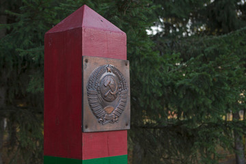 The top of wooden border post painted in green and red horizontal stripes with a metal plate depicting the coat of arms of the USSR attached in its top on the background of green coniferous trees