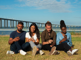 Diverse team of friends obsessed with gadgets. Focused young men and women sitting on grass, using smart phones. Addiction concept