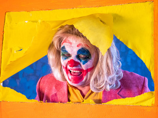 Portrait of male clown in pink dressed for Halloween. actor made makeup from crazy evil clown, faced with bloody smile. White face makeup. Colour wig. Model in Halloween costume rips yellow paper