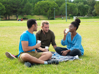 Diverse group of friends enjoying coffee break in park. Young men and woman sitting around plaid on grass, drinking takeaway coffee and chatting. Friends meeting concept