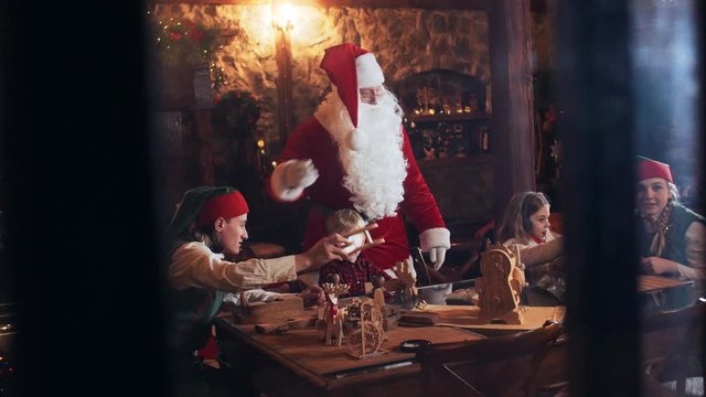 Santa Claus and elves are playing with children with wooden toys while sitting at a table in the New Year's residence. View from the street through the window