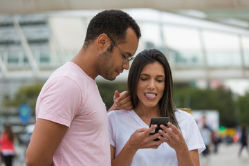 Smiling good friends looking at smartphone on street. Cheerful multiracial couple using smartphone. Communication and technology concept