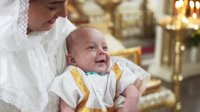 Happy caucasian mother with scarf on head is holding her adorable smiling 3-months son in russian church