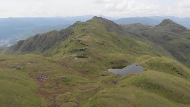 An aerial forward footage of a grassy mountain plateau rocky summit with small pond and mountain range in the background under a cloudy sky