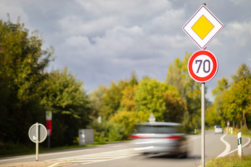 A yellow square sign on the main road and a round red and white sign with a speed limit of 70 kilometers per hour on a German country road. A car drives past high speed.