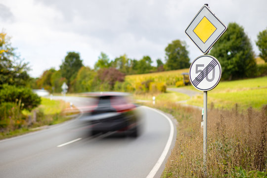 A yellow square sign on the main road and a round white sign. End of the speed limit of 50 kilometers per hour on a German highway. A car drives past high speed.