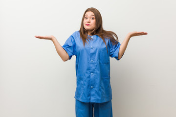 Young nurse woman against a white wall confused and doubtful shrugging shoulders to hold a copy...