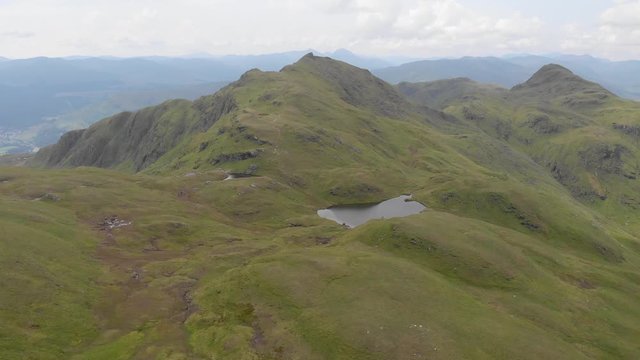 An aerial sideway left to right footage of a grassy mountain plateau rocky summit with small pond and mountain range in the background under a cloudy sky