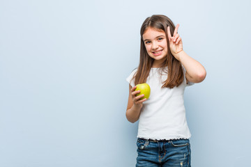 Little caucasian girl holding a green apple showing victory sign and smiling broadly.