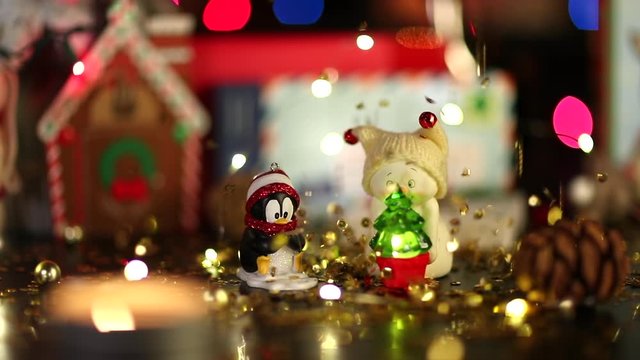 A festive still life video of Christmas decor under the rain of gold sequins. Сute penguin toy and little snowman with a small fir are in the foreground. Defocused lights at the background.