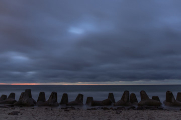 dramatic gloomy landscape of the sea coast with concrete breakwaters on the beach under a dusky gray cloudy sky at long exposure