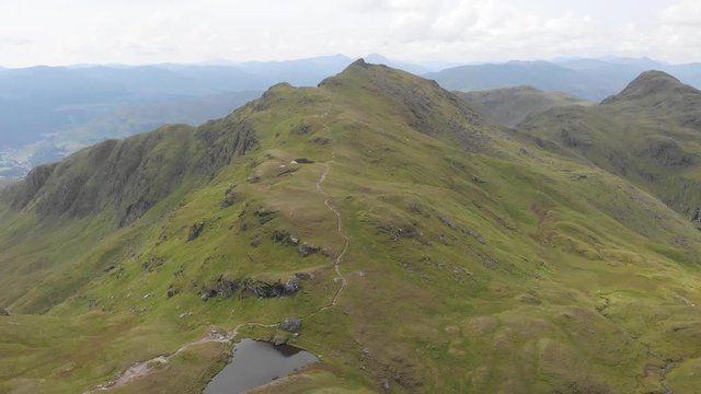 An aerial backward footage of a grassy mountain plateau rocky summit with small pond and mountain range in the background under a cloudy sky
